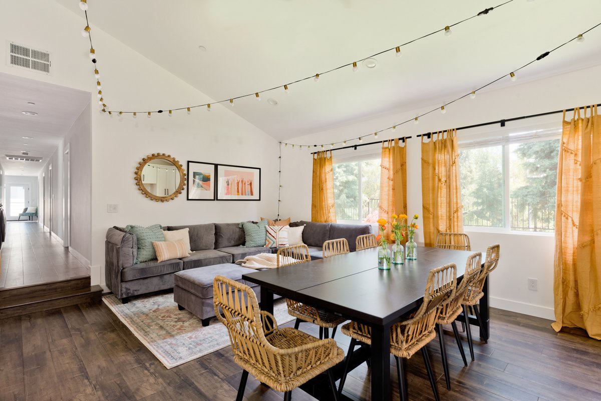 Dining table that seats 8 in the middle of the room. Bright yellow curtains drape the windows. There is a large gray L-shaped couch in the back with square two ottoman stools in front
