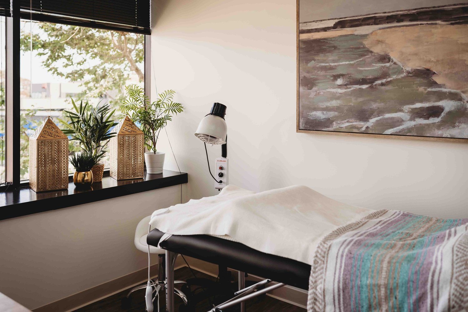 Acupuncture Therapy, a clean white room with plants and large framed art on the wall. There's a medical bed with white and colorful sheets on it which a lamp hovering above the bed