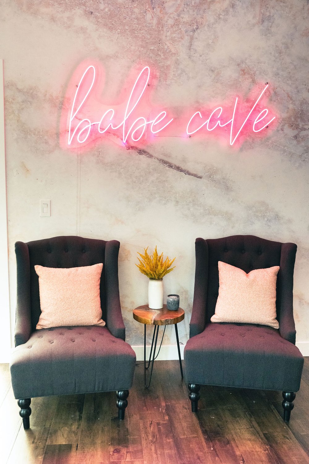 pink LED sign that says 'babe cave' hangs on the wall above two identical gray arm chairs with a small wooden stool table between them