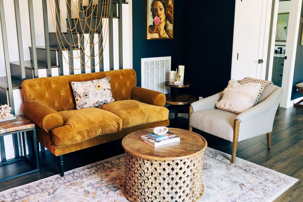 Sitting area: Light brown velvet loveseat and gray armchair on the side with a round wicker table in front. In the back are stairs with wooden slats supporting it