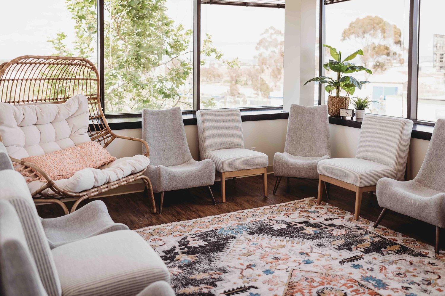 Group Therapy Room #1; many gray and white chairs and 1 wide brown wicker chair positioned in a circle in a well lit, sunny room. There is a large colorful rug on the floor