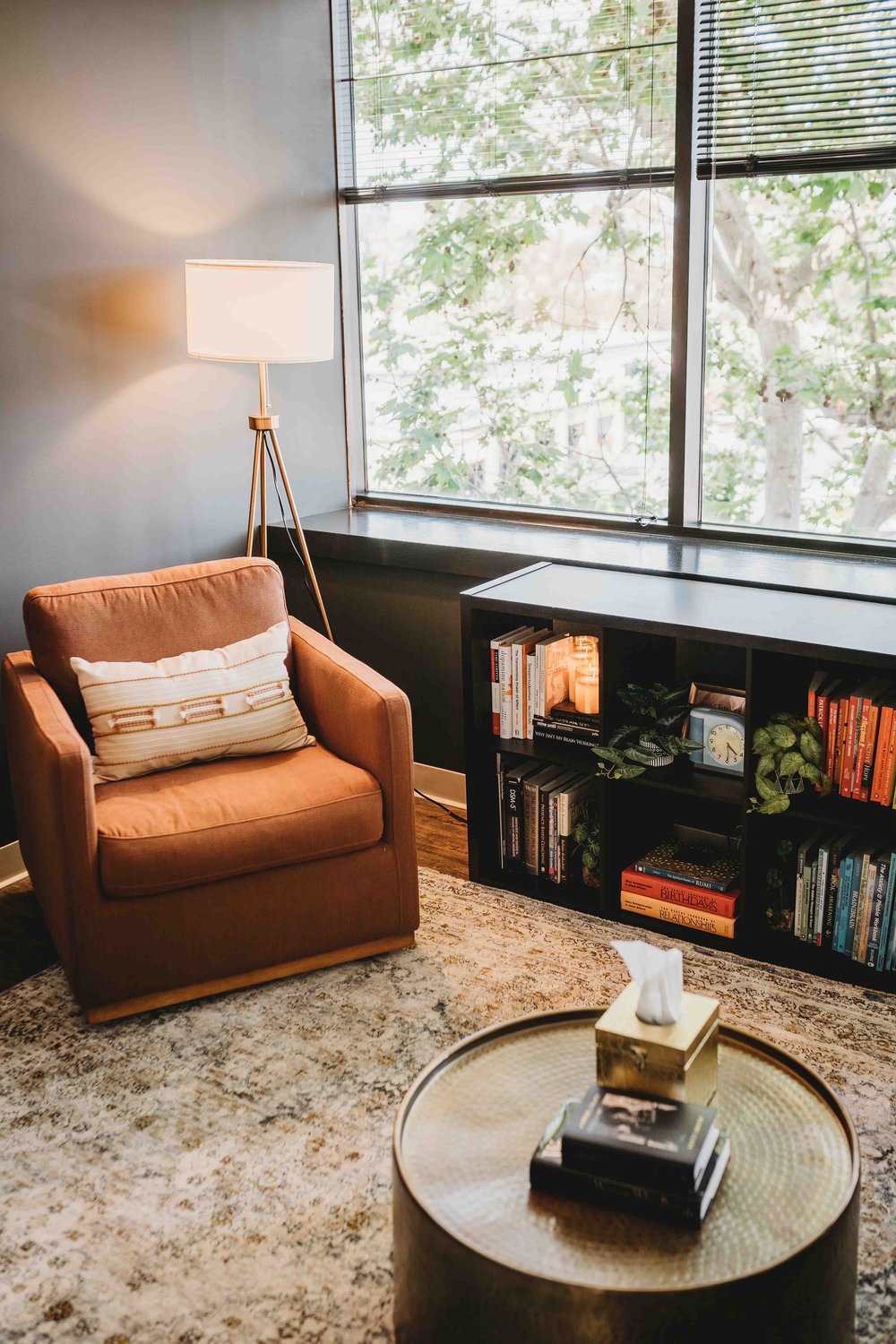 Individual Therapy Room: Room with large windows, a single-seating orange couch with a pillow on it, low 2-row book shelves, a round table with a tissue box and a stack of books