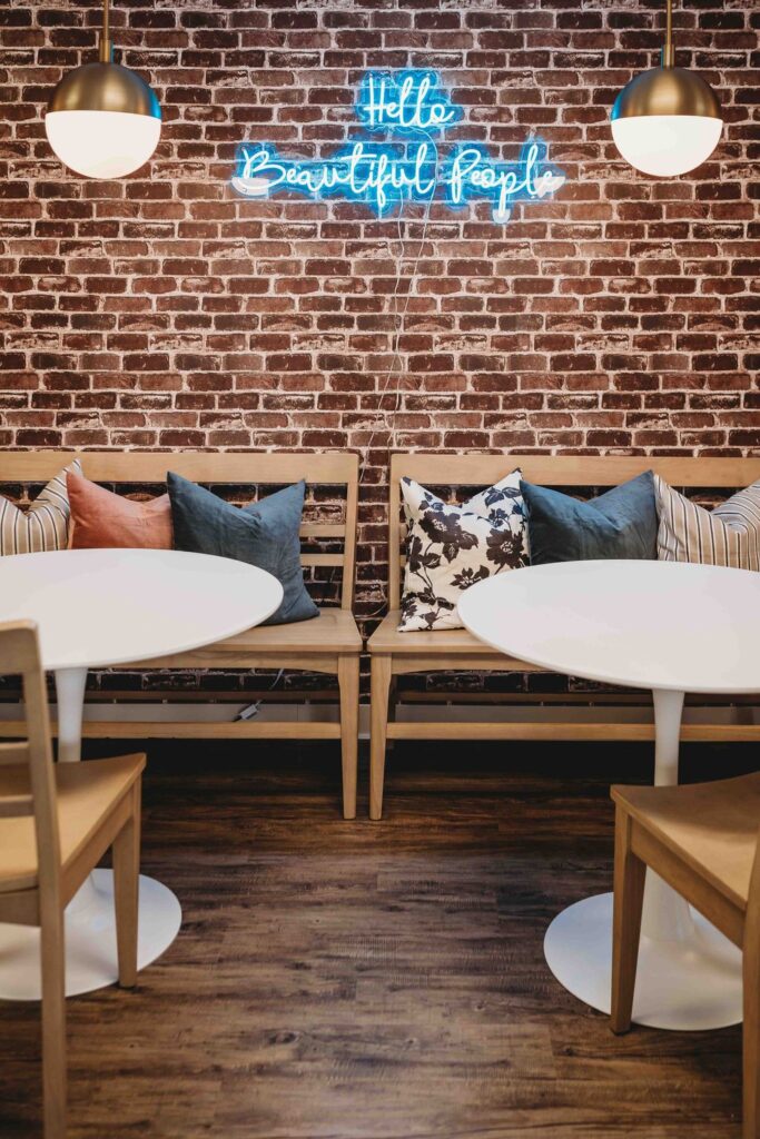 Cafe; a blue LED sign that says 'Hello Beautiful People' hang on a brick wall. There are two identical ceiling lights, round white tables, and wooden benches with pillows on them