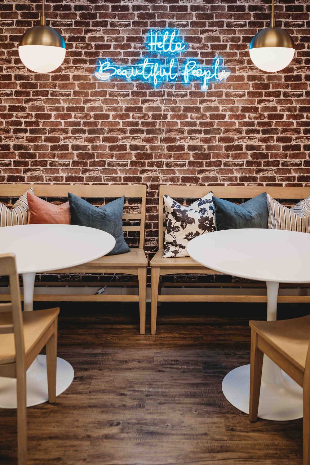 Cafe; a blue LED sign that says 'Hello Beautiful People' hang on a brick wall. There are two identical ceiling lights, round white tables, and wooden benches with pillows on them