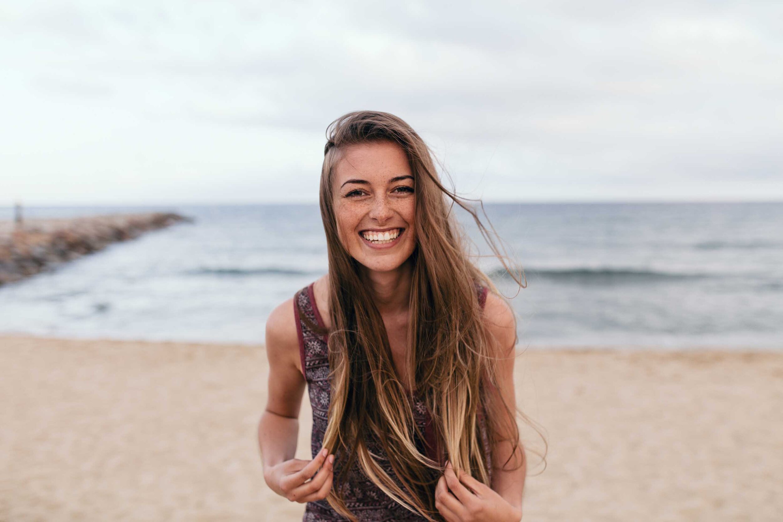Long haired woman with freckles smiles at the camera on the beach with the ocean behind her