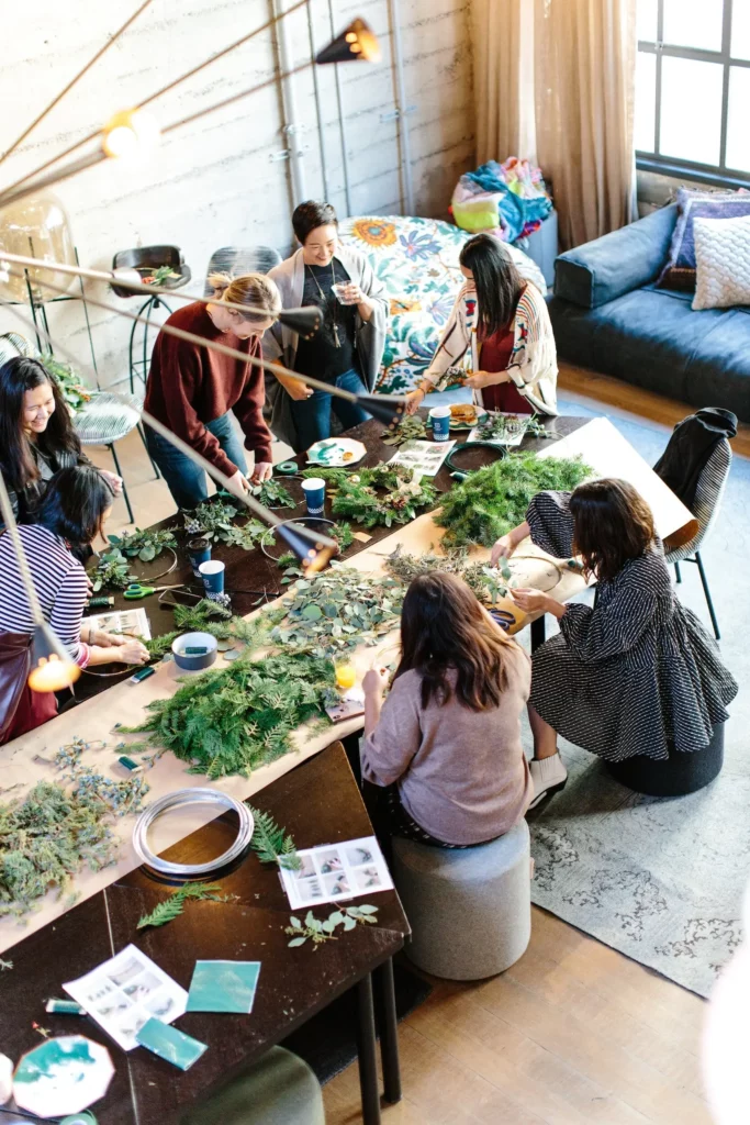 Group of women gathered around a long wooden table doing a crafts activity. Making wreathes out of different leaves and foliage