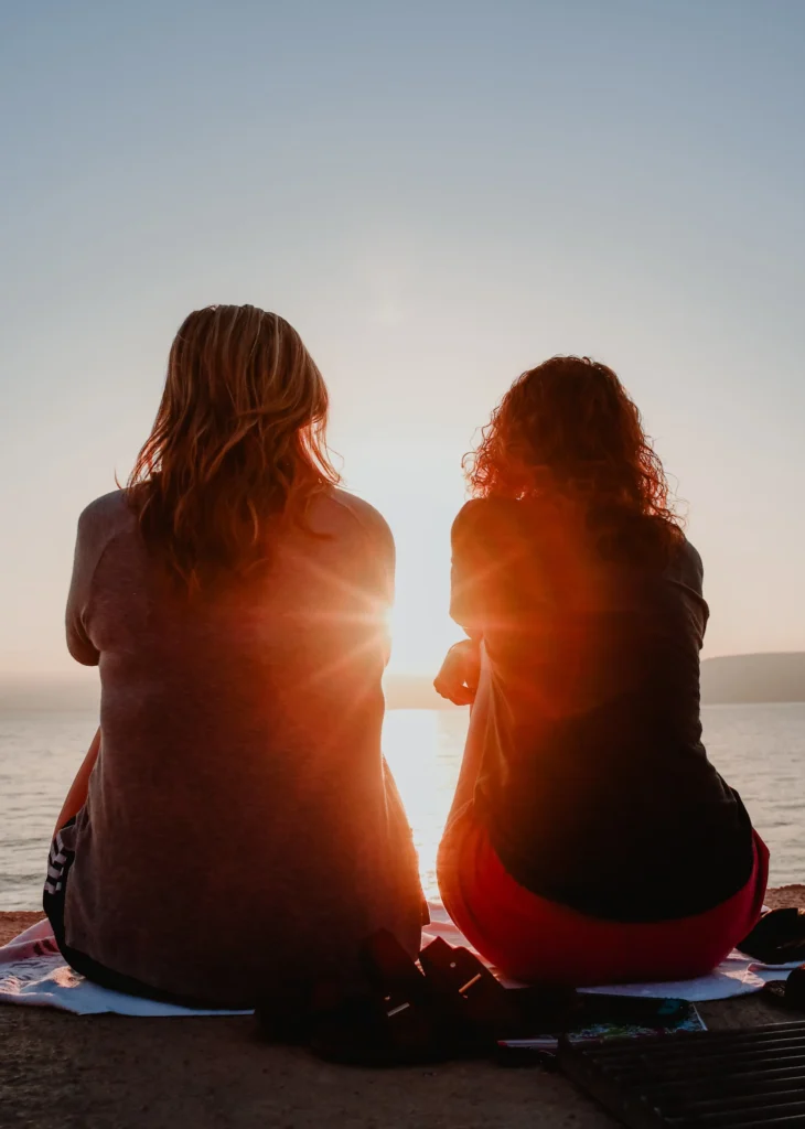 Two women sitting on a log on the beach looking out towards the sunset/sunrise over the ocean