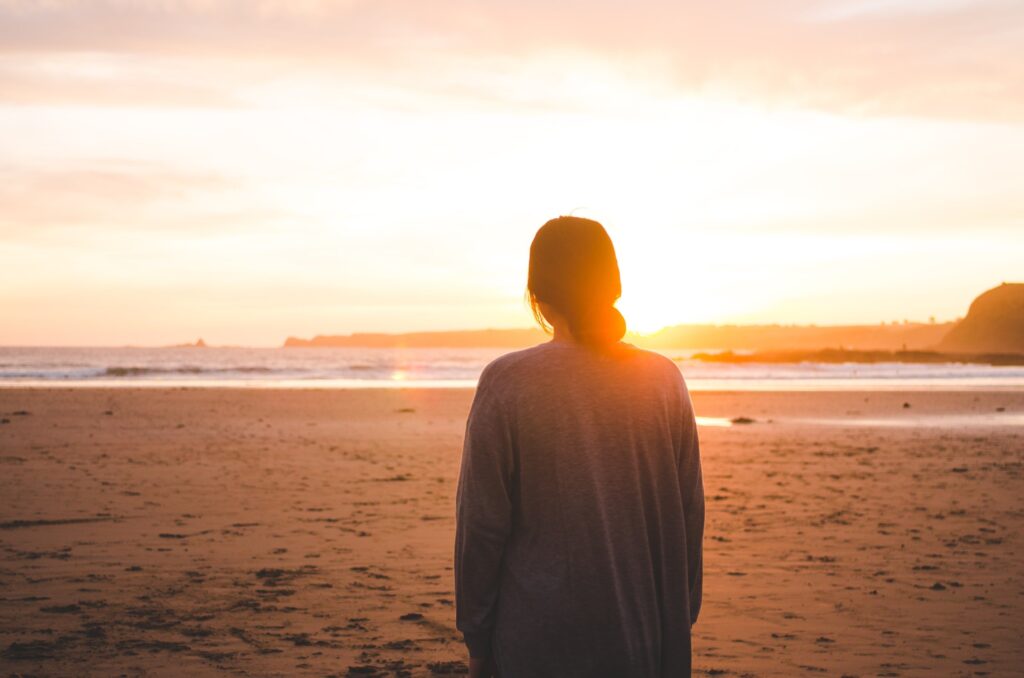 1 woman with her hair in a low bun and gray sweater looking towards the ocean at sunset
