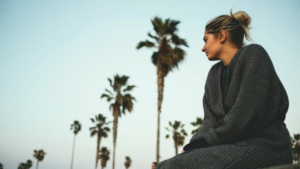 Woman with her hair in a bun sitting on a ledge looking off in the distance. There are rows of palm trees reaching towards the sky behind her