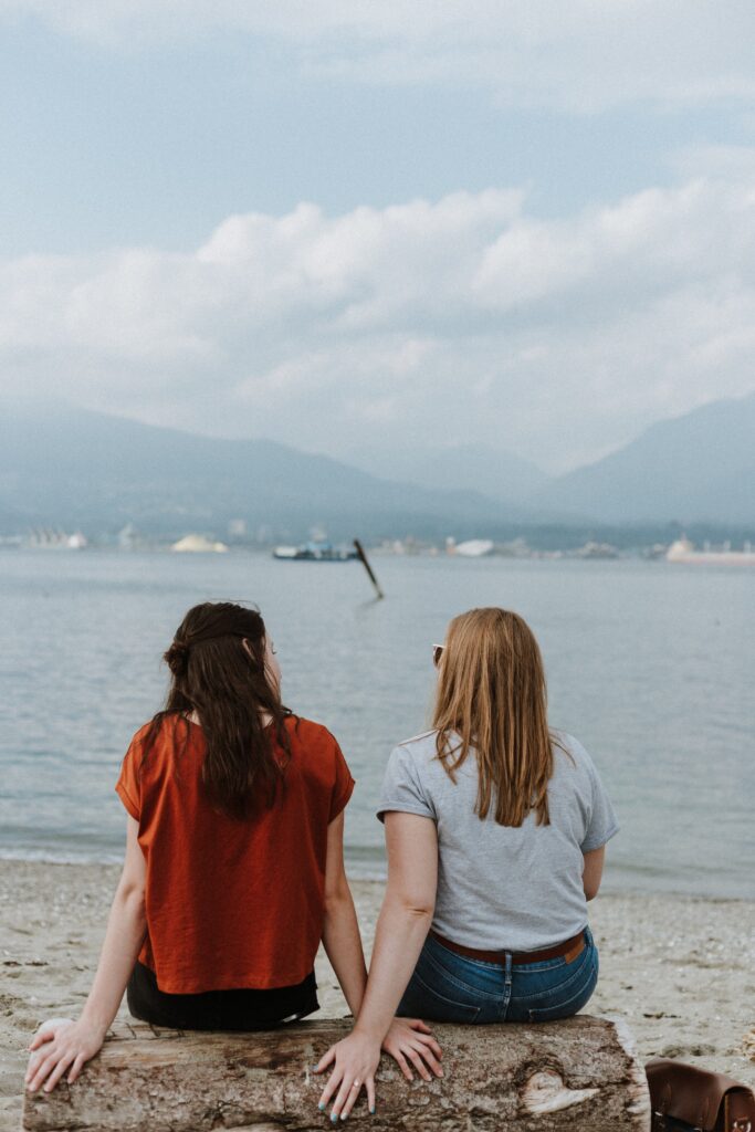 Two women sitting on a wooden log on the beach and talking while looking out at the ocean