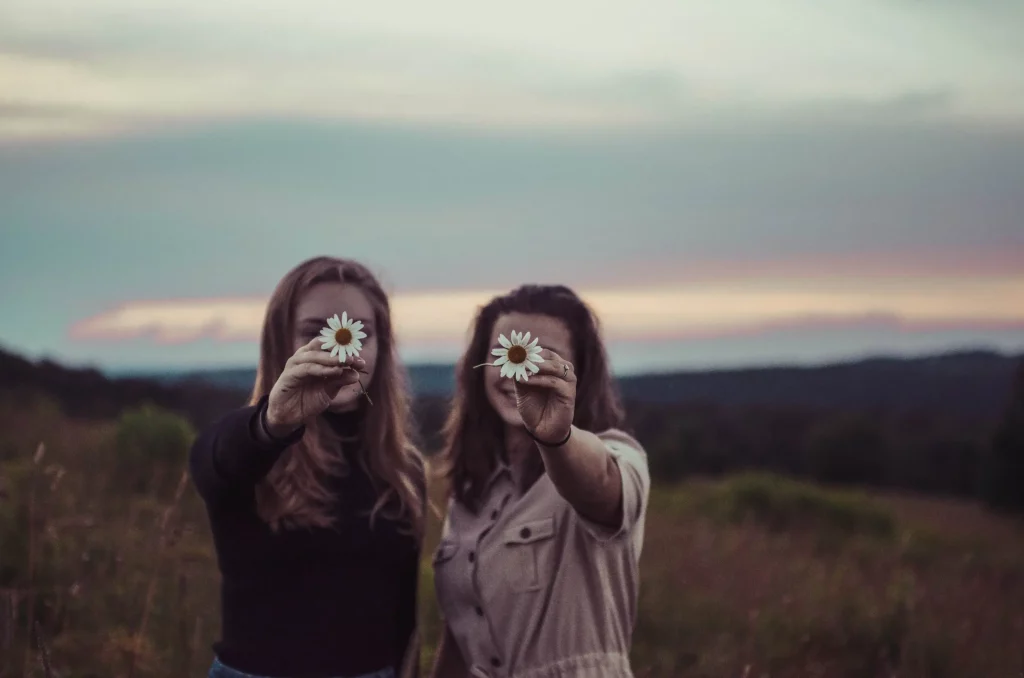 Two women holding white daisies towards the camera. The daisies are covering their faces. They stand in a grassy empty field