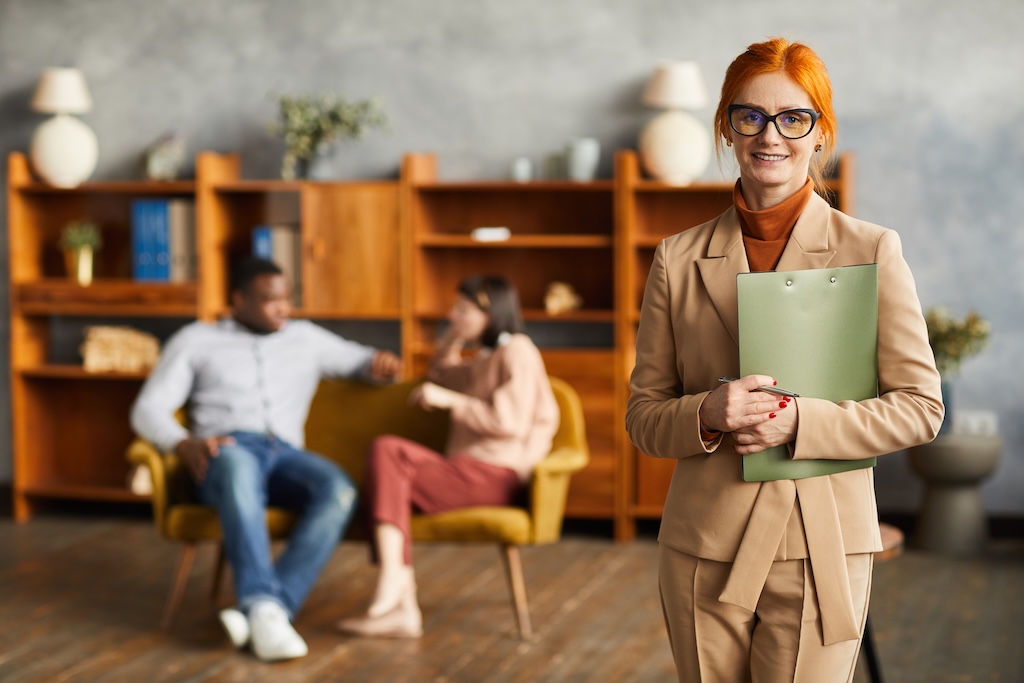 Portrait of successful psychologist standing with documents and smiling at camera with couple sitting in the background