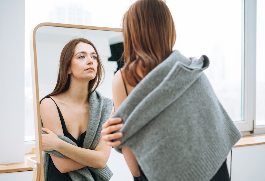 Young woman with long dark hair on black dress with gray sweater looking in mirror