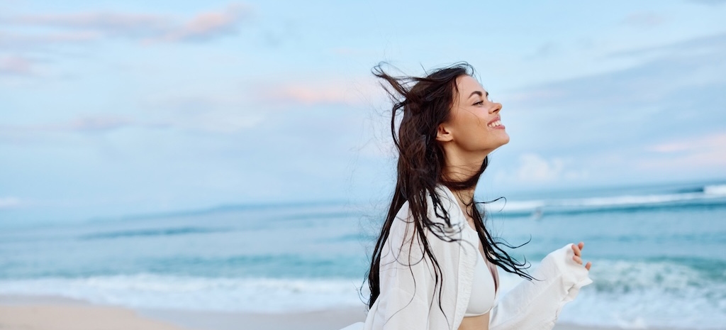 Brunette woman with long hair in a white shirt and denim smiling on the beach