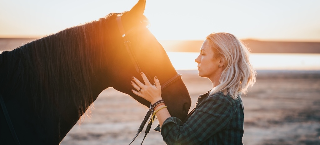 Blonde woman stroking and hugging horse at sunset