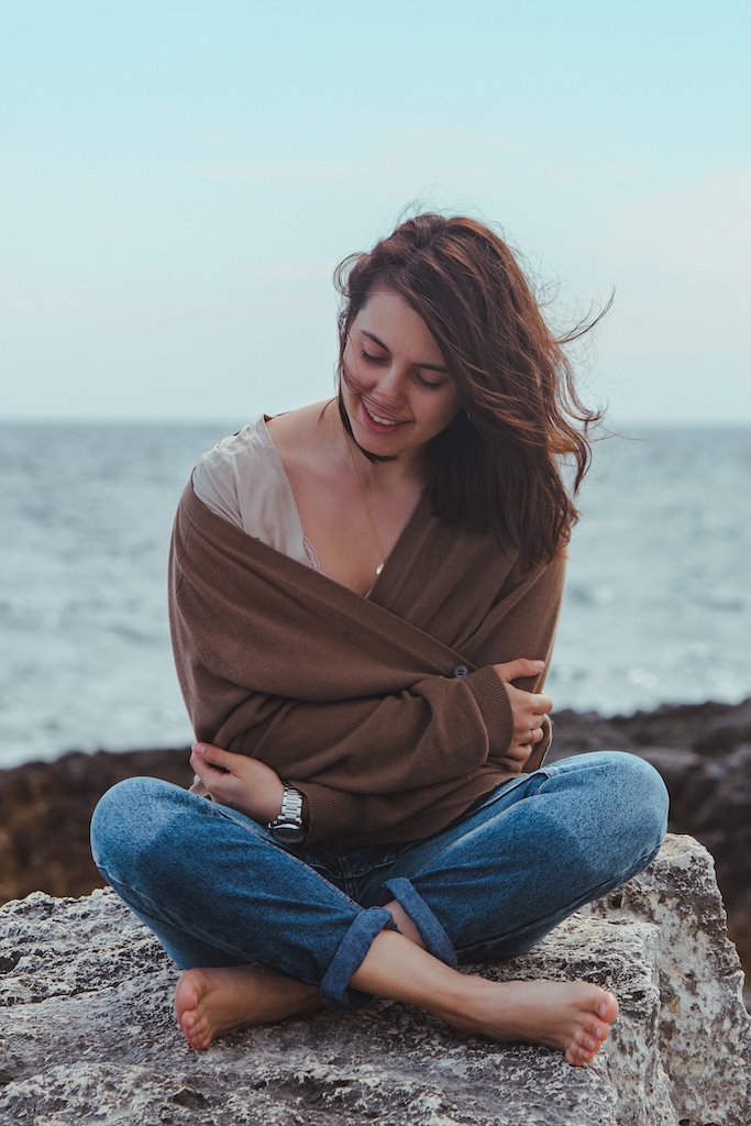woman sitting by rocky sea beach in wet jeans lighthouse on background. windy weather.