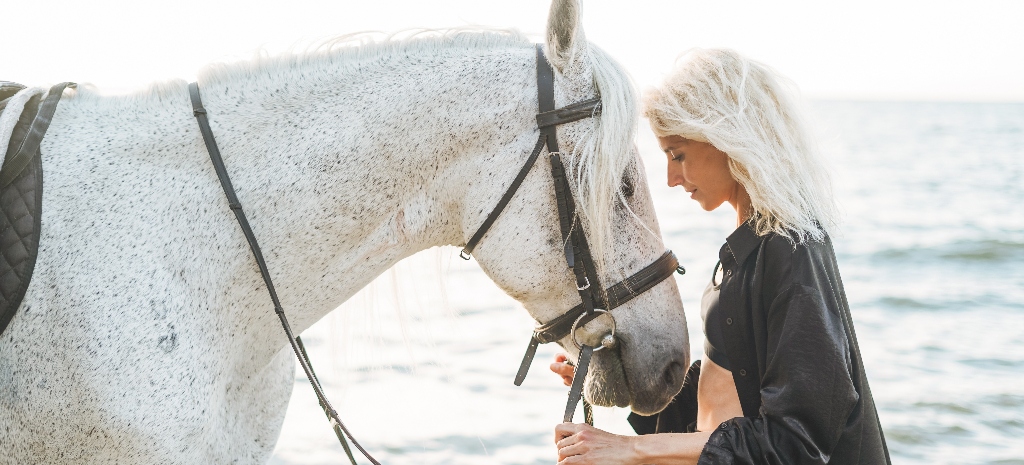 Young blonde woman in black clothes riding white horse on seascape background