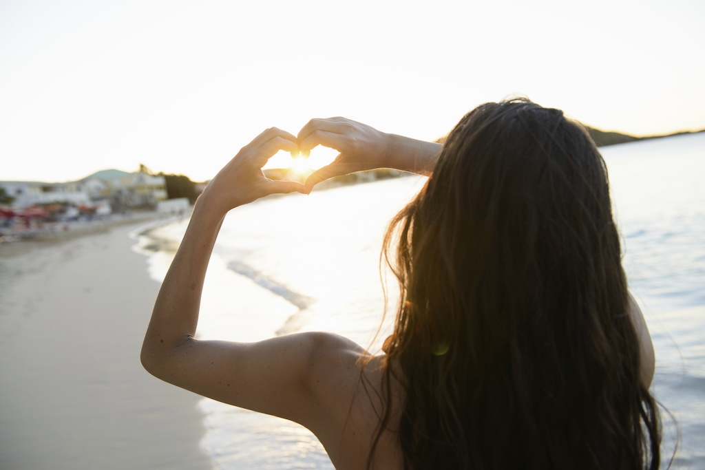 Woman making heart shape around sun with her hands on the beach