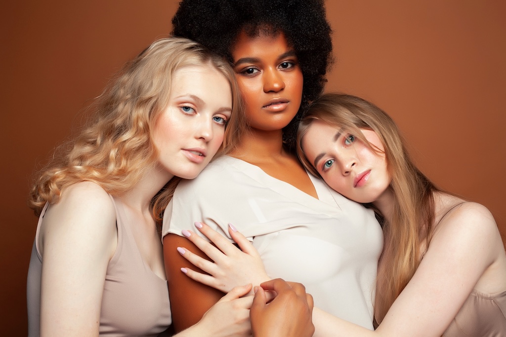 three diverse women, supporting and hugging each other with solid brown background, representing women's healing with trauma therapy, which we offer at Monima Wellness