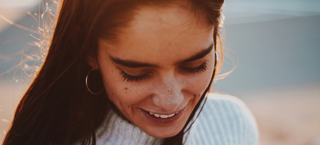 Woman with dark hair and features looking down at the ground, hair blowing in the wind, at the beach. Representing healing and freedom from addiction.