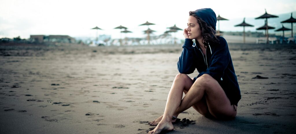 Young woman sitting on the beach, dark skies, depicting struggles with mental health or substance use