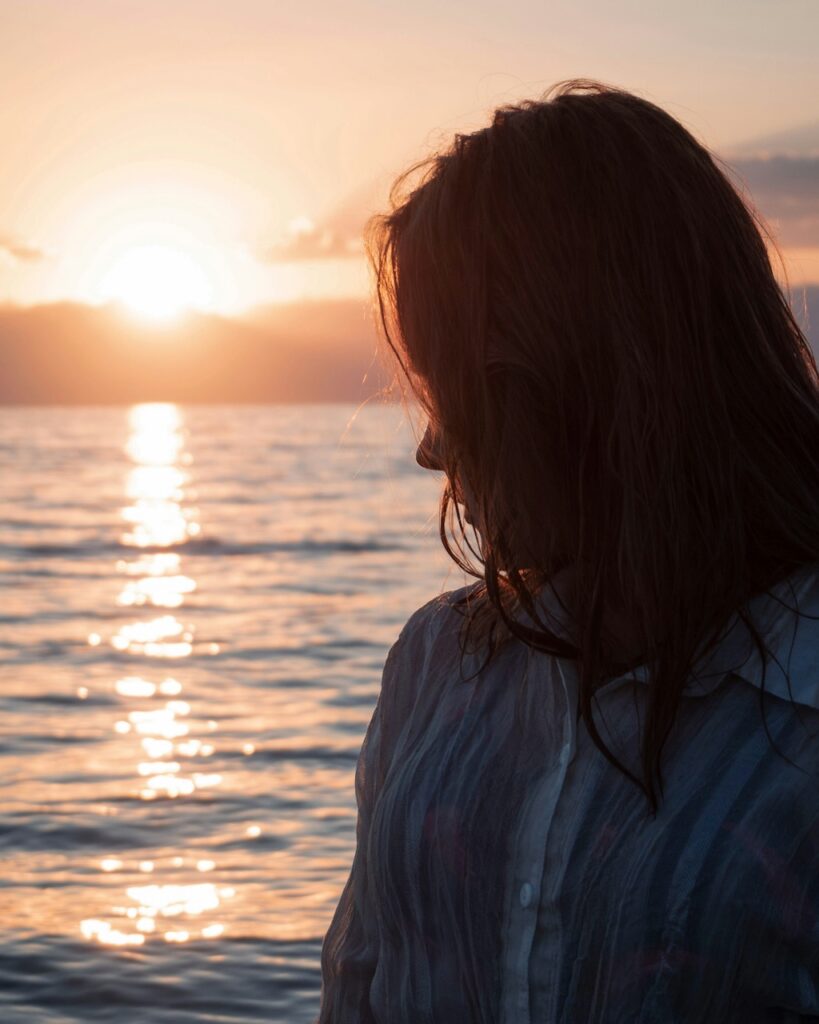 Woman looking down toward the ocean at sunset in soft, warm light, reflecting a quiet, introspective moment and the emotional impact of substance use challenges