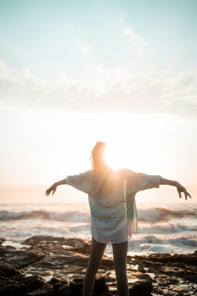 Woman standing at the ocean during sunset with arms open in soft, warm light, representing a sense of relief, hope, and support through ADHD treatment and integrated mental health care