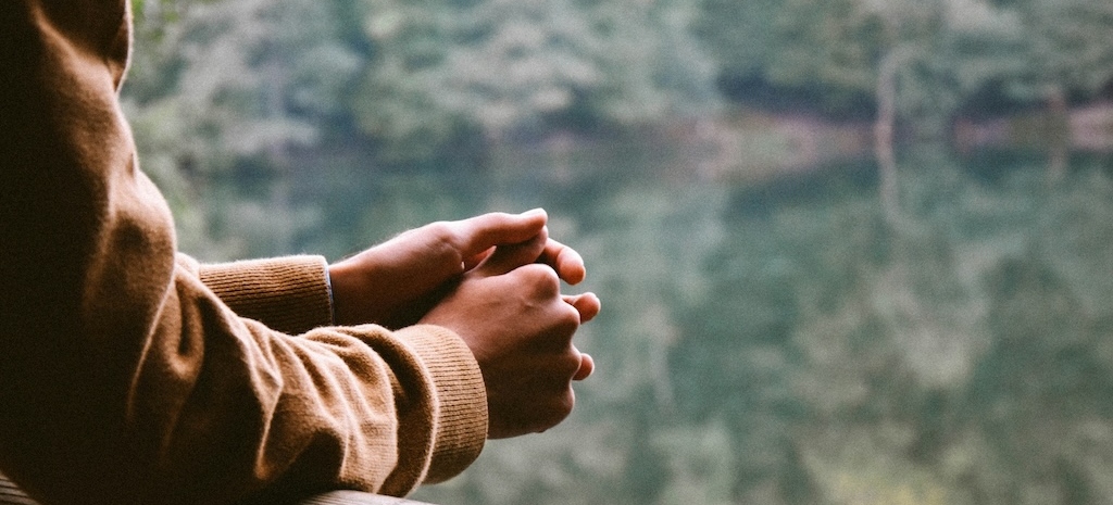 A person in a warm sweater resting their clasped hands on a railing, gazing out over a calm, tree-lined lake—evoking stillness, grounding, and quiet reflection.