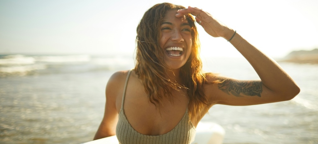 Woman smiling and holding a surfboard at the beach, reflecting a sense of empowerment and forward movement in beginning the healing process from trauma