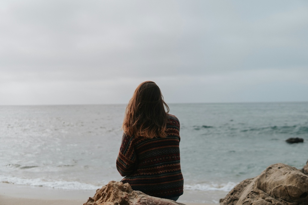 Person sitting quietly on coastal rocks facing the ocean, reflecting a calm and supportive environment for sexual trauma treatment at Monima Wellness Center