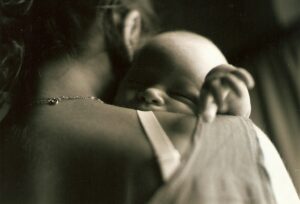 Black and white close-up of a mother holding a sleeping baby against her shoulder, conveying tenderness, comfort, and the emotional experience of early postpartum motherhood.