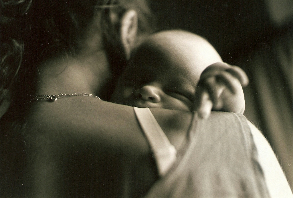 Black and white close-up of a mother holding a sleeping baby against her shoulder, conveying tenderness, comfort, and the emotional experience of early postpartum motherhood.