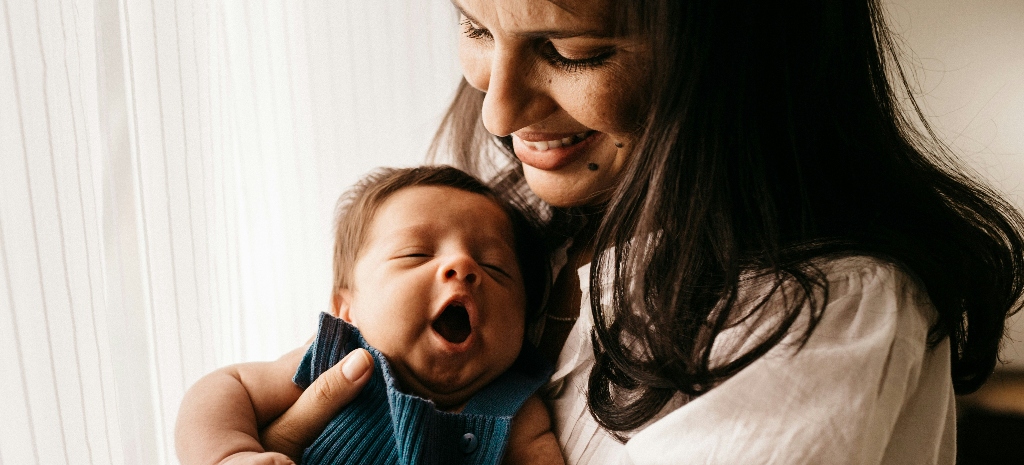 Mother holding a yawning newborn near a window with soft natural light, representing the emotional connection and care involved in the postpartum experience.