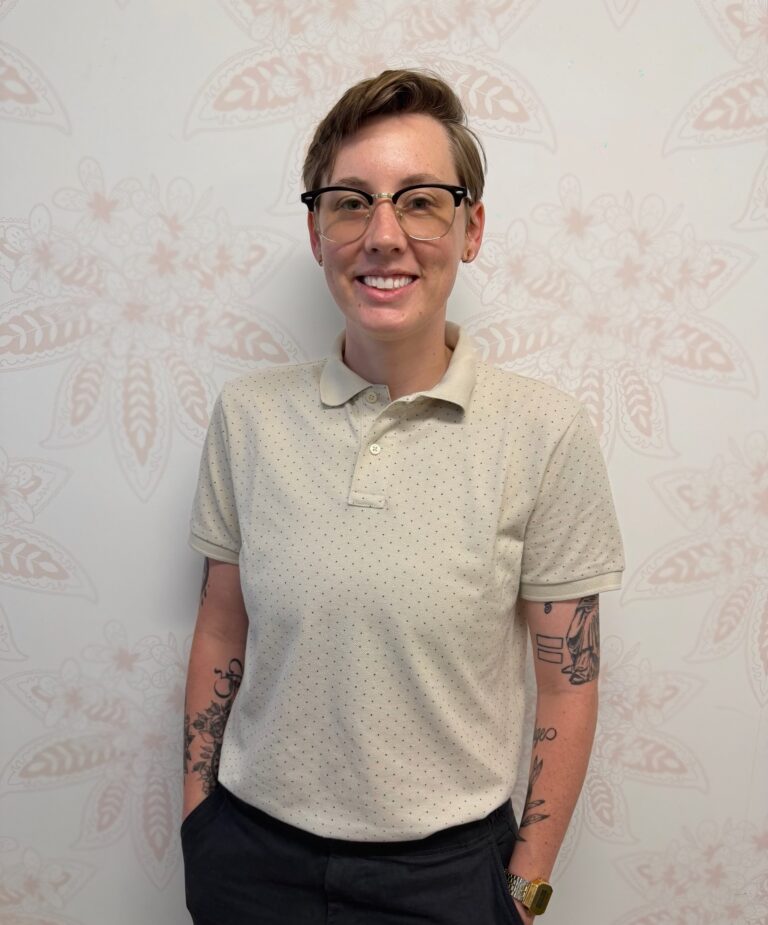 Linzy Holmes, Admissions Coordinator at Monima Wellness Center, standing and smiling in front of a light floral-patterned wall. They are wearing glasses, a cream-colored collared shirt, dark pants, and a gold watch, with visible tattoos on both arms.