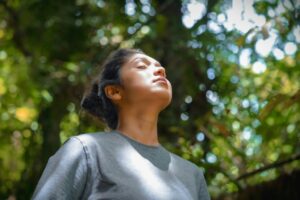 Woman taking a calming breath outdoors beneath green trees, breathing and regulating nervous system.